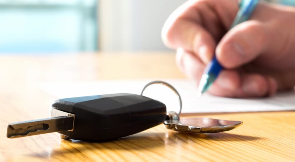 A close up of a car key and a hand filing out paperwork to sell their car.