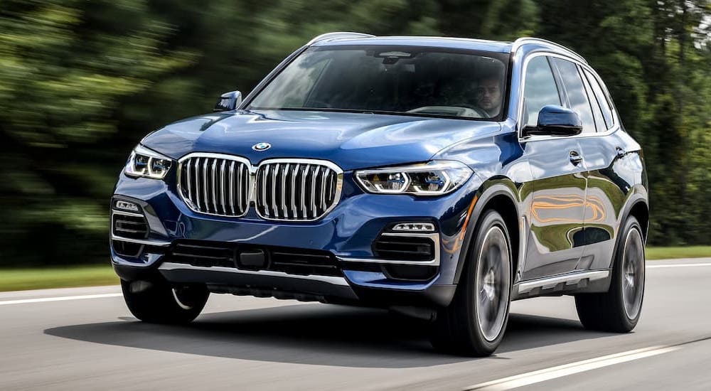A blue 2019 BMW X5 is shown driving on a tree-lined road by a used car dealer near Fishers.